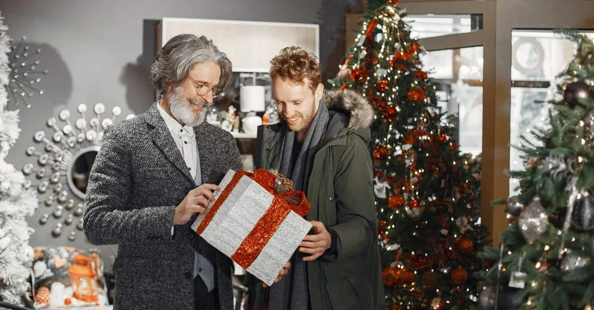 Deux hommes échangeant joyeusement un cadeau au milieu des sapins de Noël décorés.