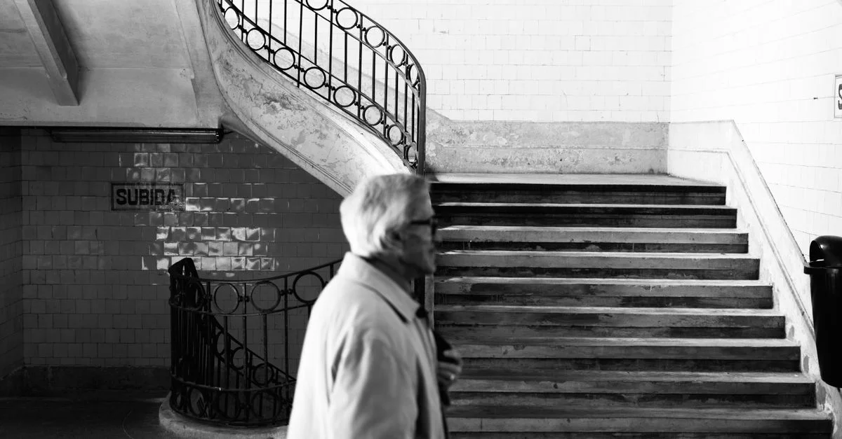 Image en noir et blanc d'un vieil homme passant devant des escaliers de métro avec des rampes élaborées.