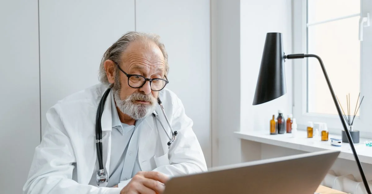 Médecin senior en blouse blanche utilisant un ordinateur portable pour un appel vidéo dans un bureau de clinique.