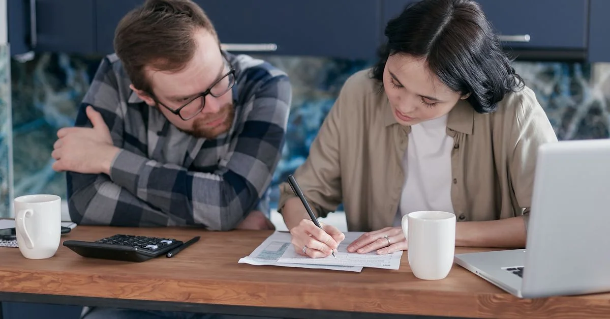 Couple adulte travaillant ensemble sur des documents à un bureau, avec un ordinateur portable et une calculatrice à proximité, dans un cadre domestique.