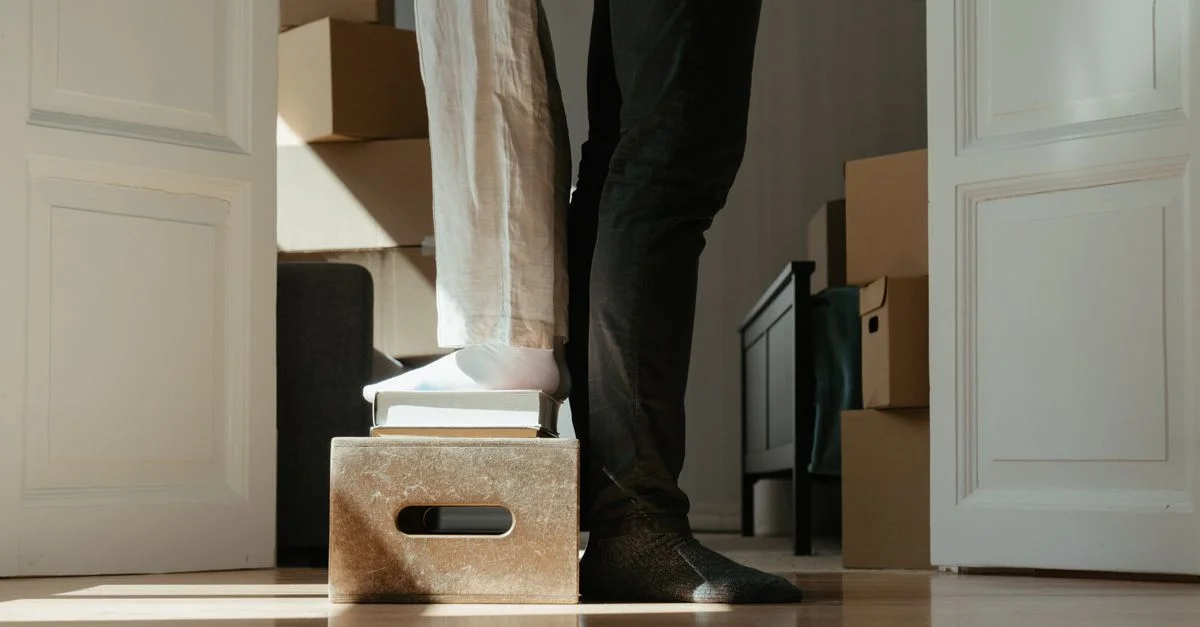 Un couple debout sur une boîte entourée de cartons en mouvement, symbolisant un déménagement.