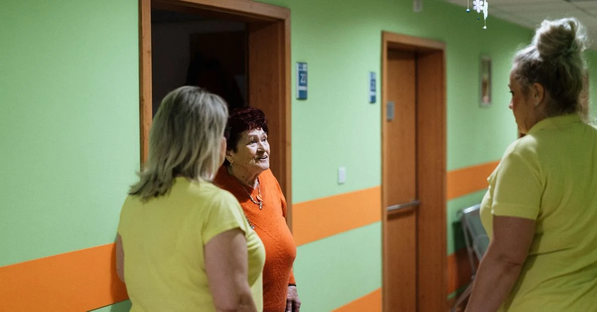 Une femme senior engage une conversation avec des aides-soignants dans le couloir d'un établissement.