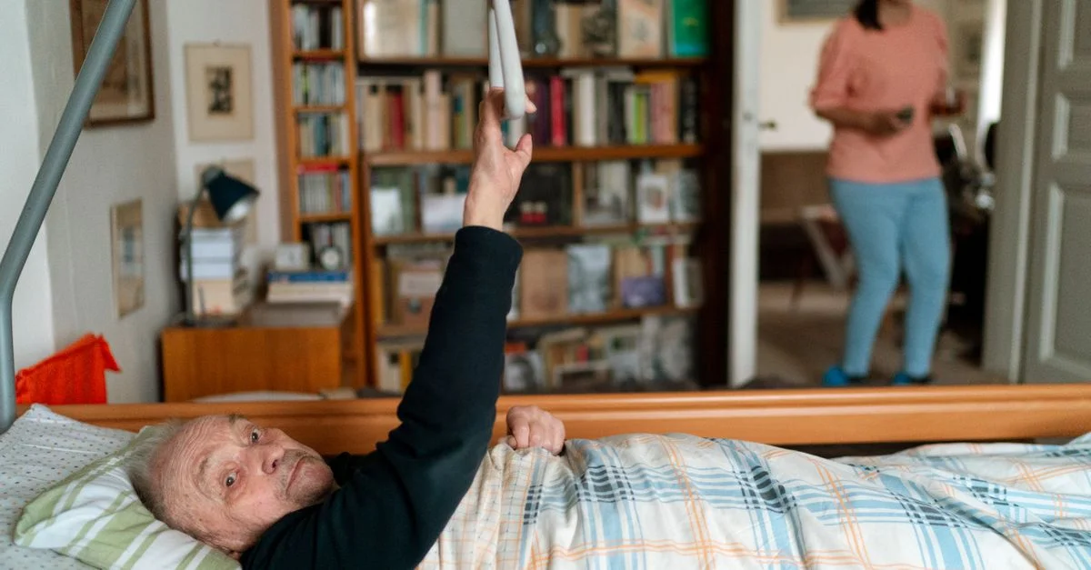 Homme âgé au lit avec un soignant dans une maison à Prague, entouré de livres.