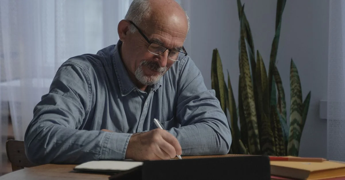 Homme senior souriant prenant des notes chez lui, incarnant la concentration et le mode de vie du travail à distance.