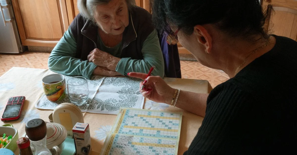 Une femme âgée et son soignant travaillent ensemble sur un jeu de mots croisés dans une cuisine chaleureuse.