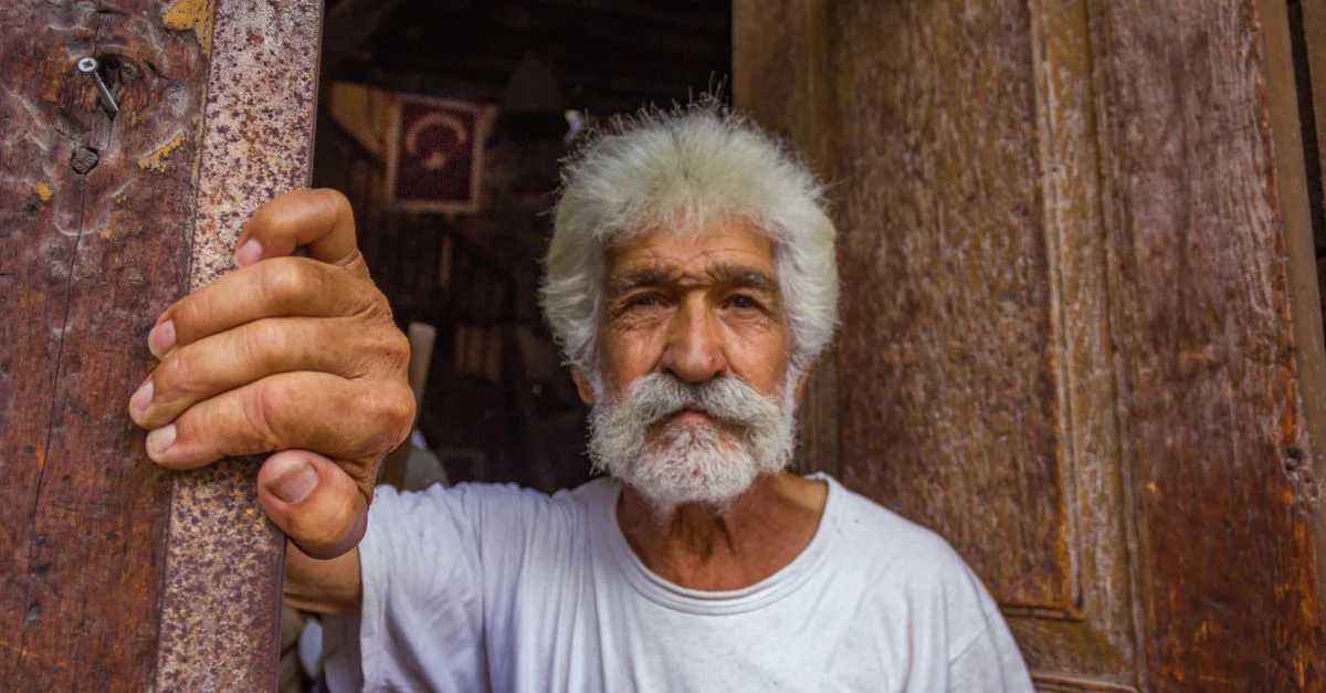 Un homme âgé avec les cheveux blancs et une barbe se tient près d'une porte en bois rustique.