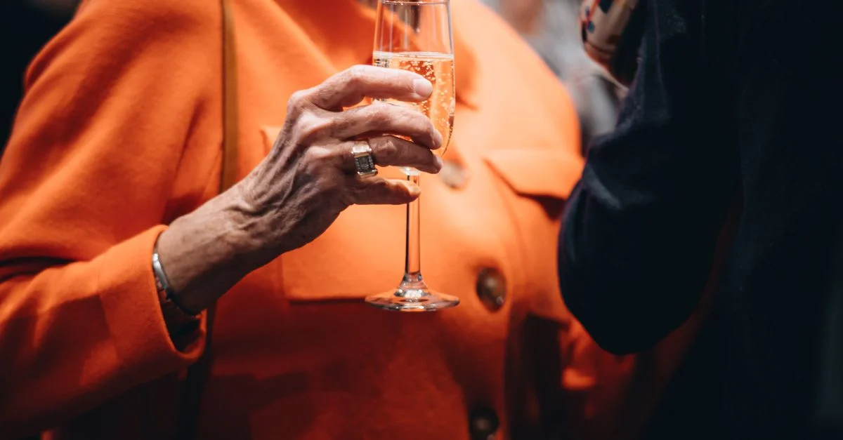Une femme âgée en manteau orange tenant une coupe de champagne lors d'un événement social, capturant l'élégance et la célébration.