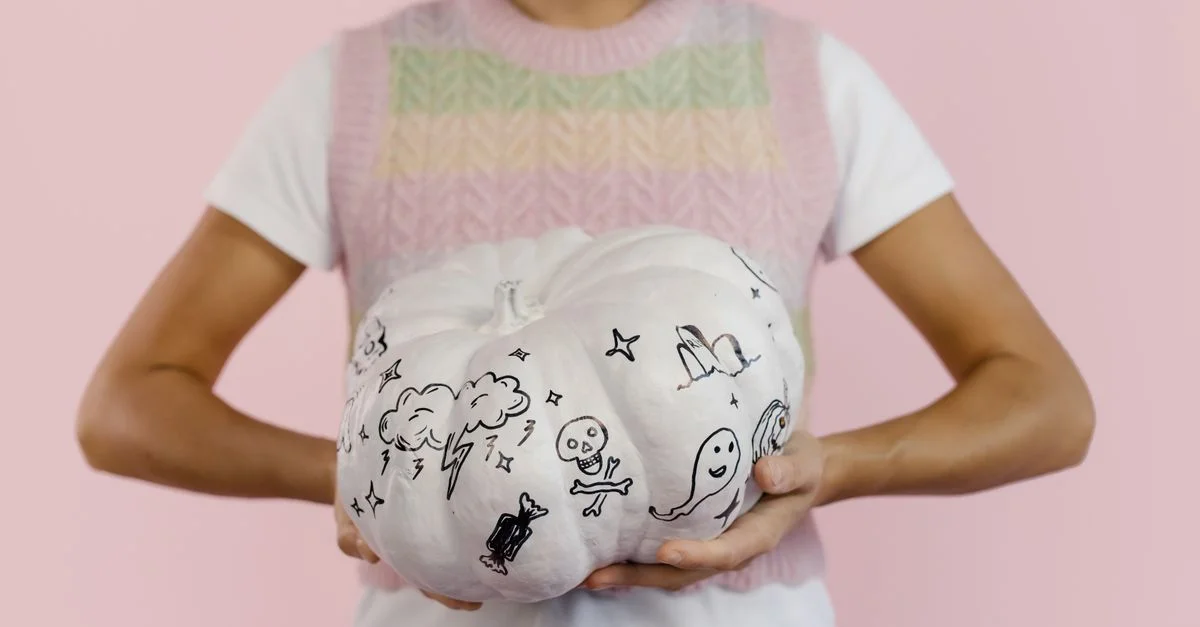Femme tenant une citrouille blanche avec des dessins effrayants sur un fond rose, idéal pour des thèmes d'Halloween.