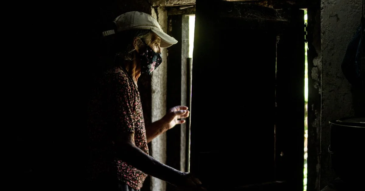 Femme senior portant un masque facial ouvre une porte dans une pièce faiblement éclairée, vue en silhouette.