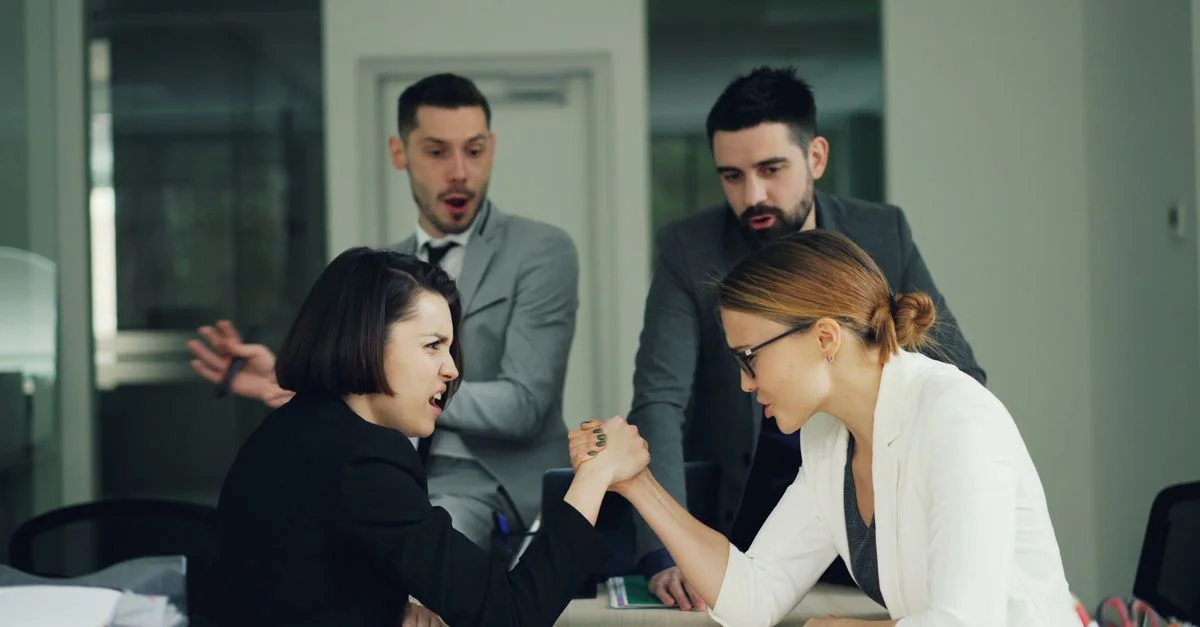 Des femmes en tenue professionnelle font un bras de fer sous le regard surpris de leurs collègues.