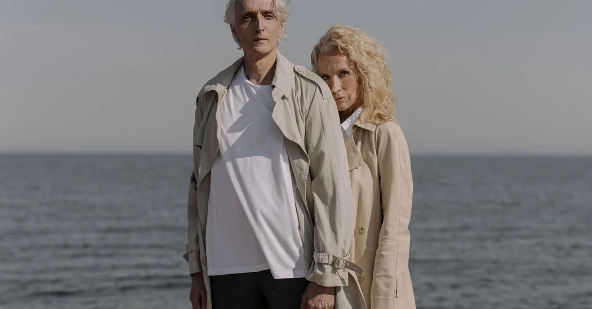 Un couple mature en trench-coats posant affectueusement sur une plage sereine, symbolisant l'amour et l'unité.