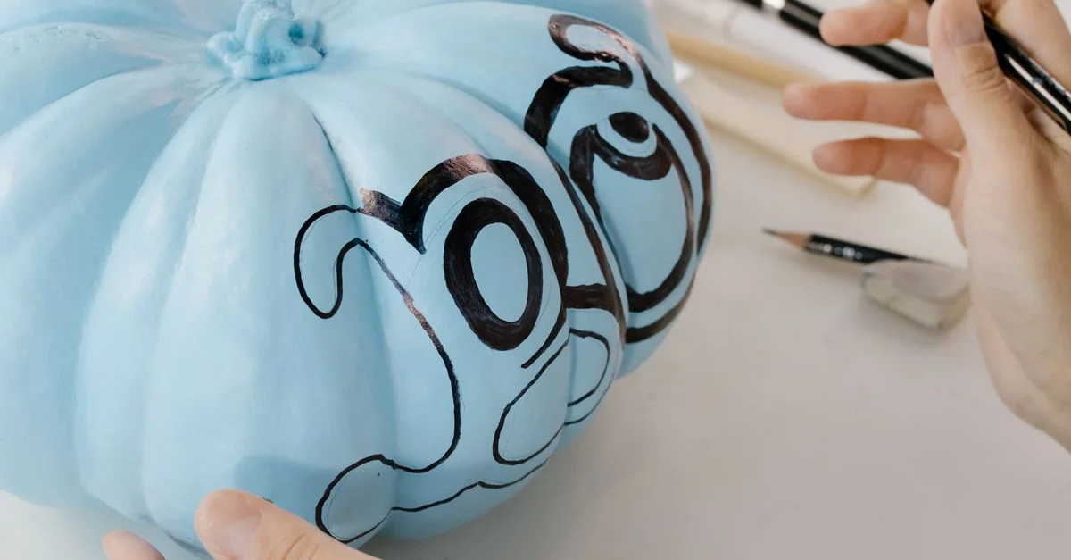 Des mains peignant une citrouille bleue avec des designs artistiques, parfaits pour des décorations créatives de Halloween.