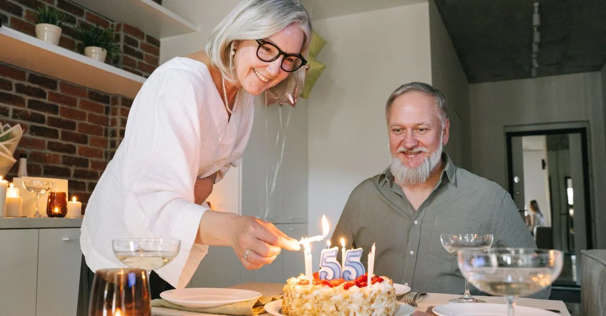 Un couple senior joyeux célèbre un 55e anniversaire avec un gâteau à la maison, entouré de bougies chaleureuses.