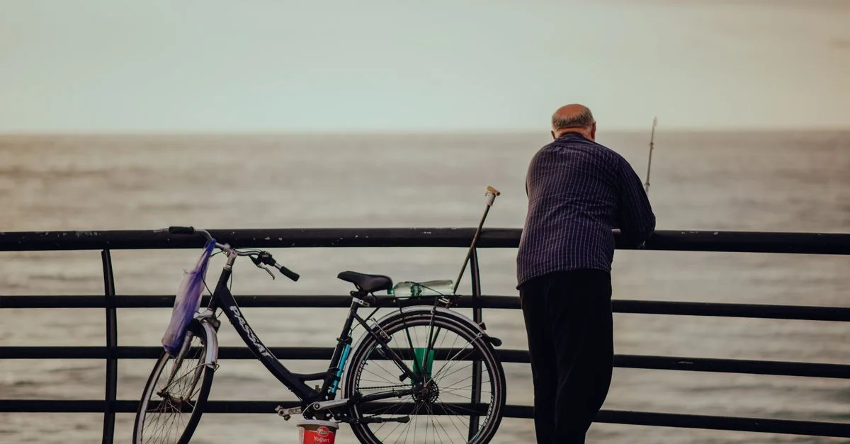 Un homme âgé pêchant depuis une jetée en bord de mer avec son vélo à proximité, offrant une scène marine paisible.