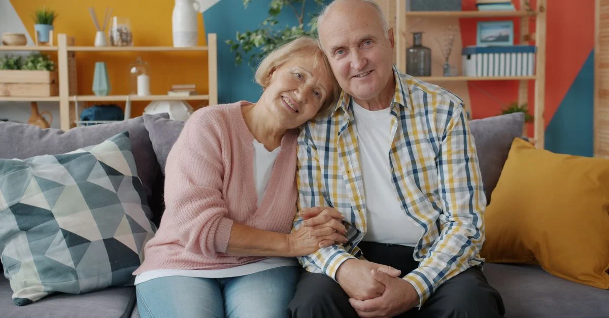 Un couple de personnes âgées souriantes, assises côte à côte sur un canapé confortable dans un salon coloré.