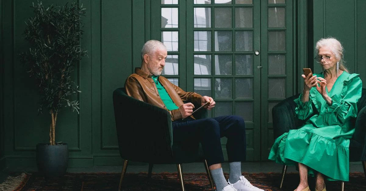 Un homme et une femme seniors utilisant des smartphones dans une pièce verte élégante avec des fauteuils et une plante.
