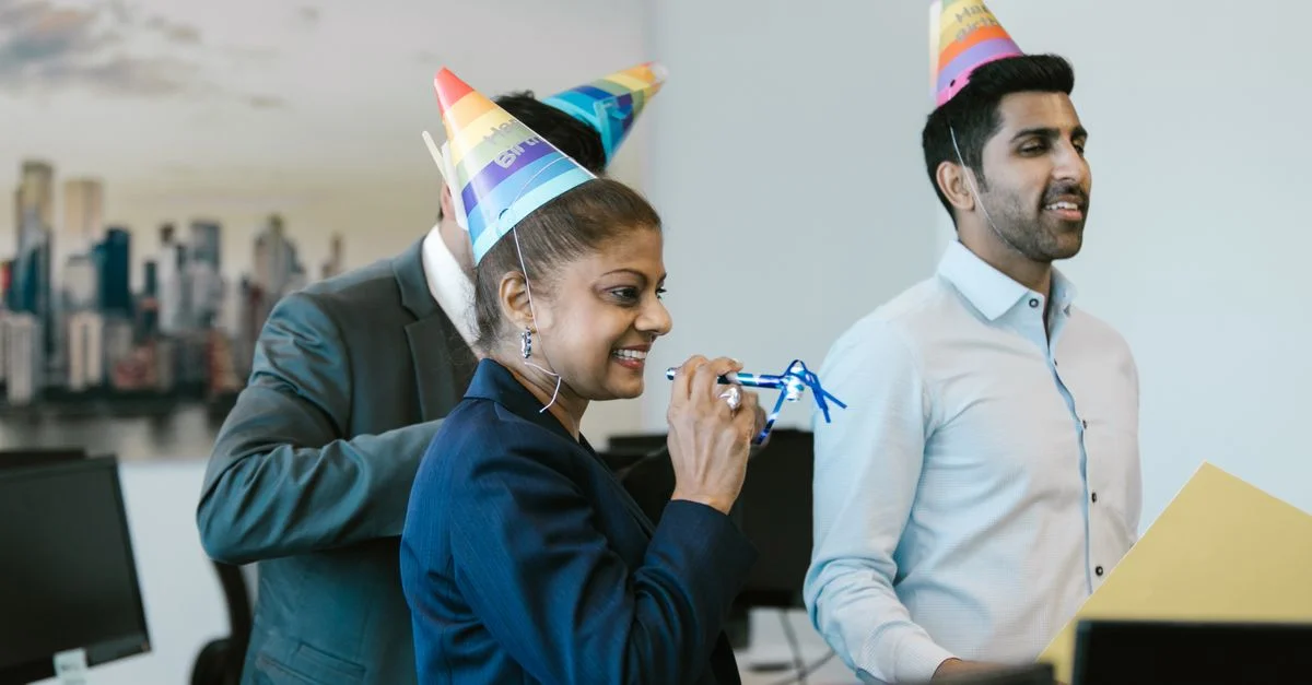 Des collègues célébrant un anniversaire au bureau avec des chapeaux colorés et des sourires.