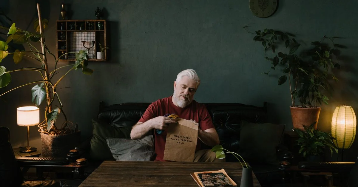 Homme senior assis dans une pièce confortable, lisant une enveloppe personnelle, entouré de plantes d'intérieur.