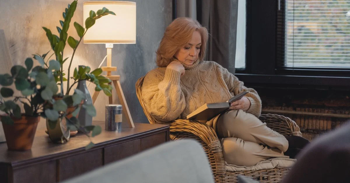 Femme senior en pull lisant dans une pièce confortable avec des plantes et une lampe.