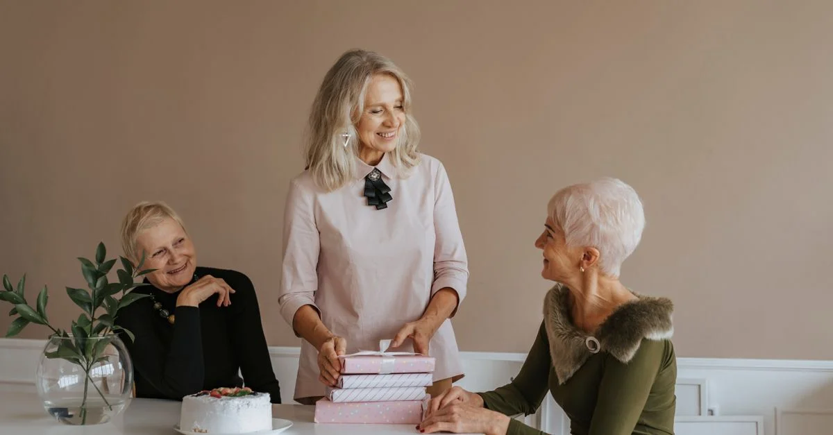 Trois femmes seniors célèbrent un anniversaire à l'intérieur avec des cadeaux et un gâteau, partageant rires et bonheur.
