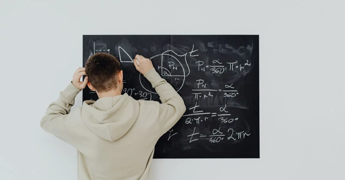Adolescent en sweat écrivant des formules mathématiques sur un tableau noir à l'intérieur, mettant en valeur ses compétences en résolution de problèmes.