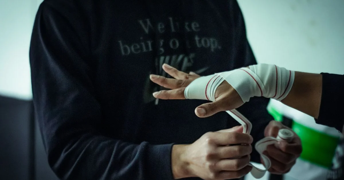 Athlète en train de s'envelopper les mains avant une séance d'entraînement dans une salle de gym faiblement éclairée, mettant en avant la préparation et la concentration.