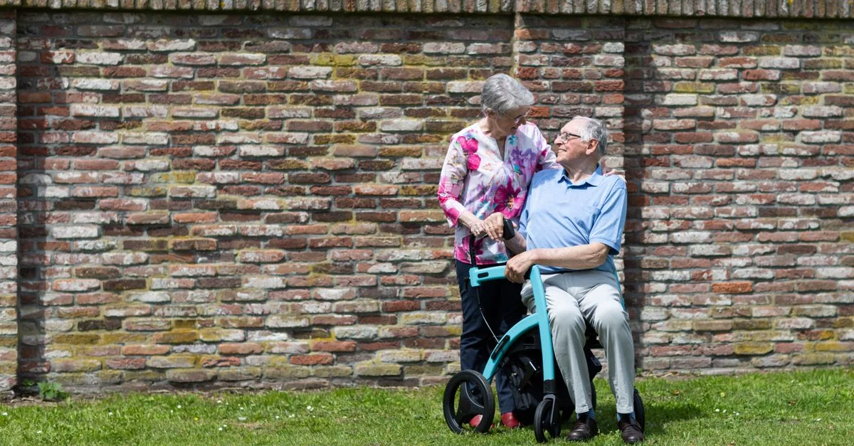 Couple âgé en extérieur, homme en fauteuil roulant assisté par une femme, exprimant amour et soin.